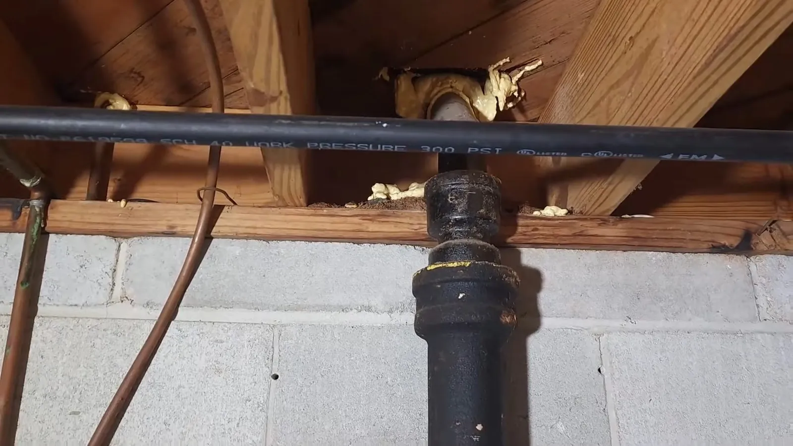 Black pipe along the floor joists above a block wall, with a black fitting and copper tubing beside it.