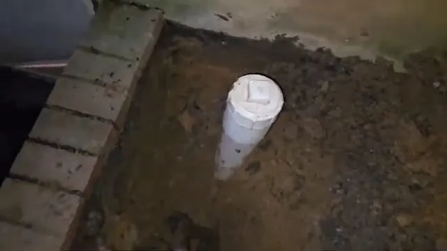 Short white vertical pipe with a cap standing in a dirt bed beside a brick wall.
