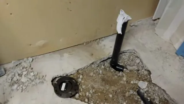 Black closet flange set in broken concrete beside a vertical drain stub with a rag in the top.