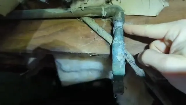 Hand pointing at a green-blue corroded copper elbow under a floor joist in a crawl space.