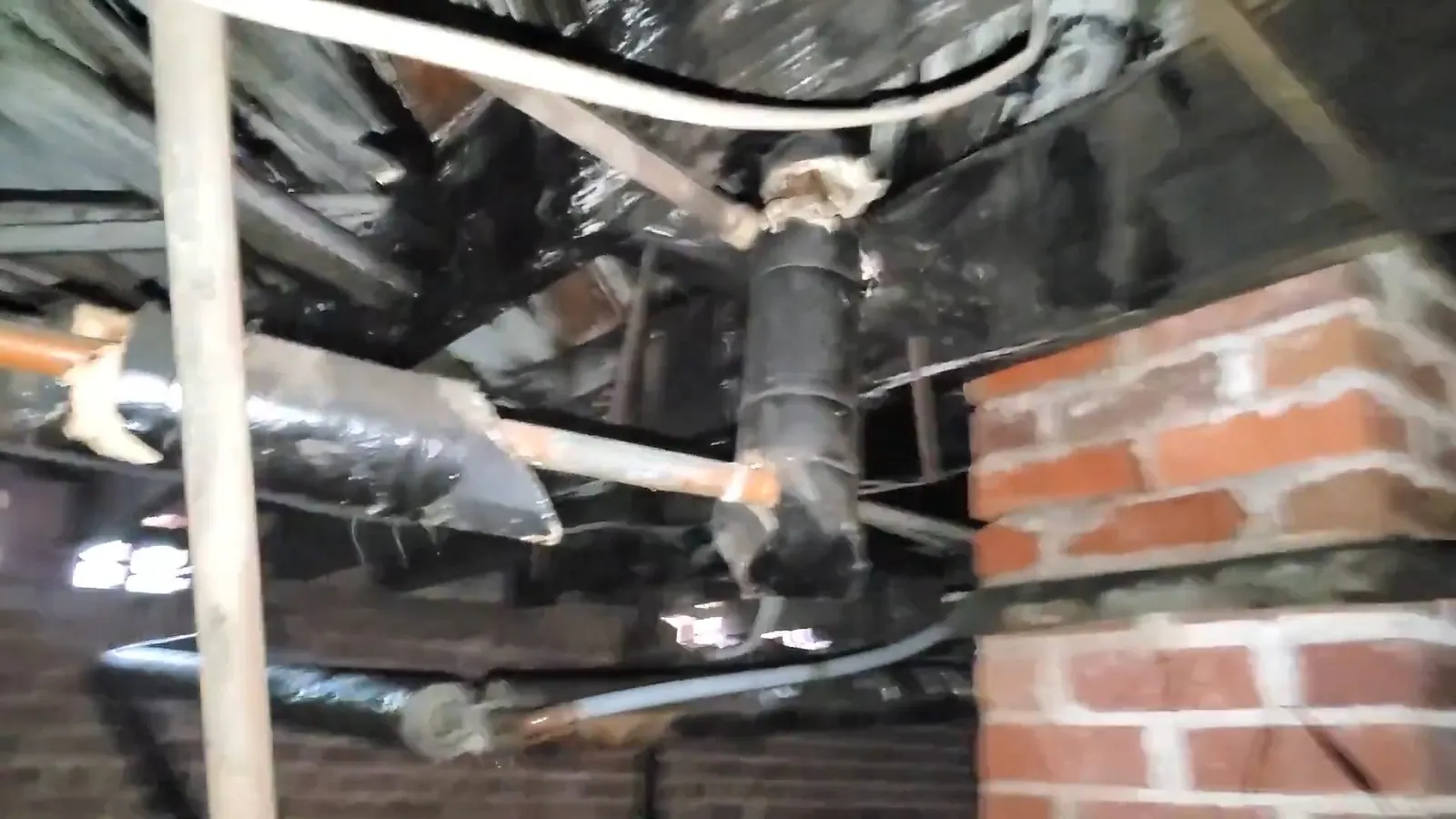 Pipes and insulated lines running under a floor beside a red brick foundation pier in a crawl space.