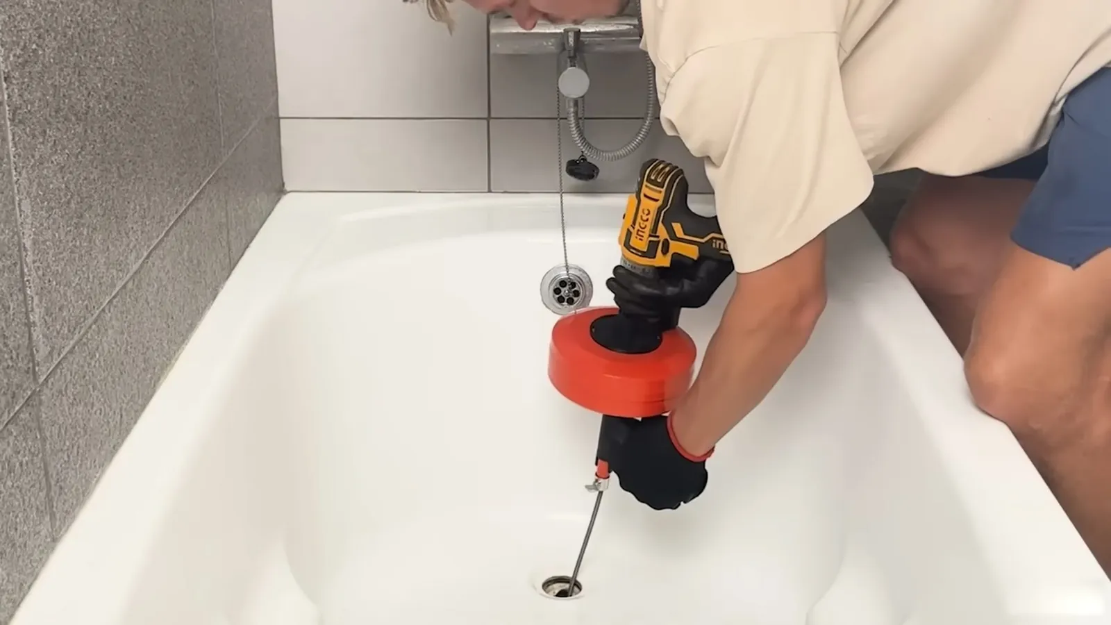 Man kneeling at a white bathtub using a drill-driven drain auger with its cable running into the drain.