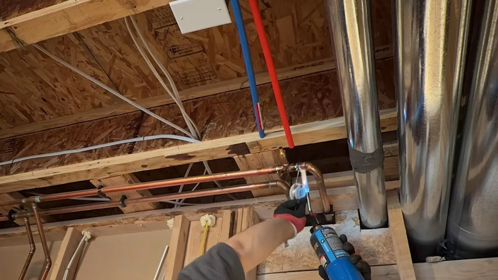 Man using a drill beside red and blue PEX tubing and copper lines between floor joists.