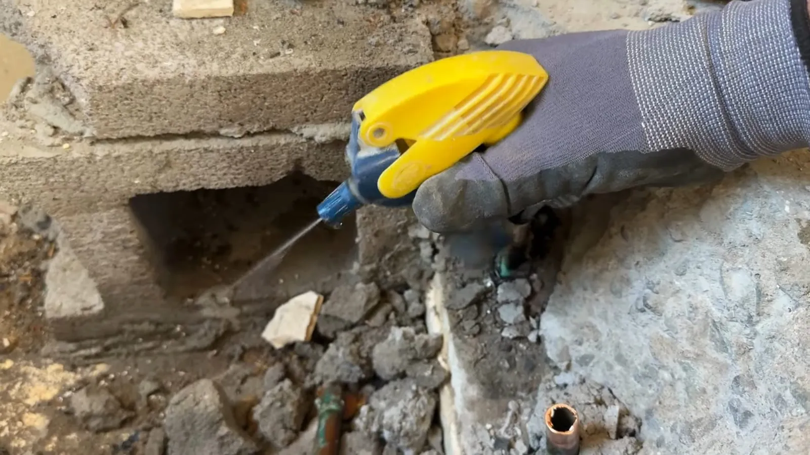Gloved hand holding a yellow epoxy applicator beside a hole broken in concrete with a copper pipe visible.