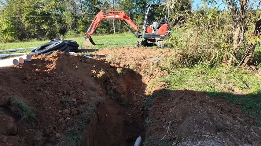 Mini excavator beside a long open trench and coiled black pipe on a grassy rural lot.