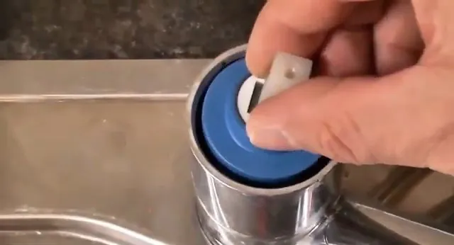 Hand lifting a blue plastic cartridge out of a chrome faucet body over a stainless steel sink.