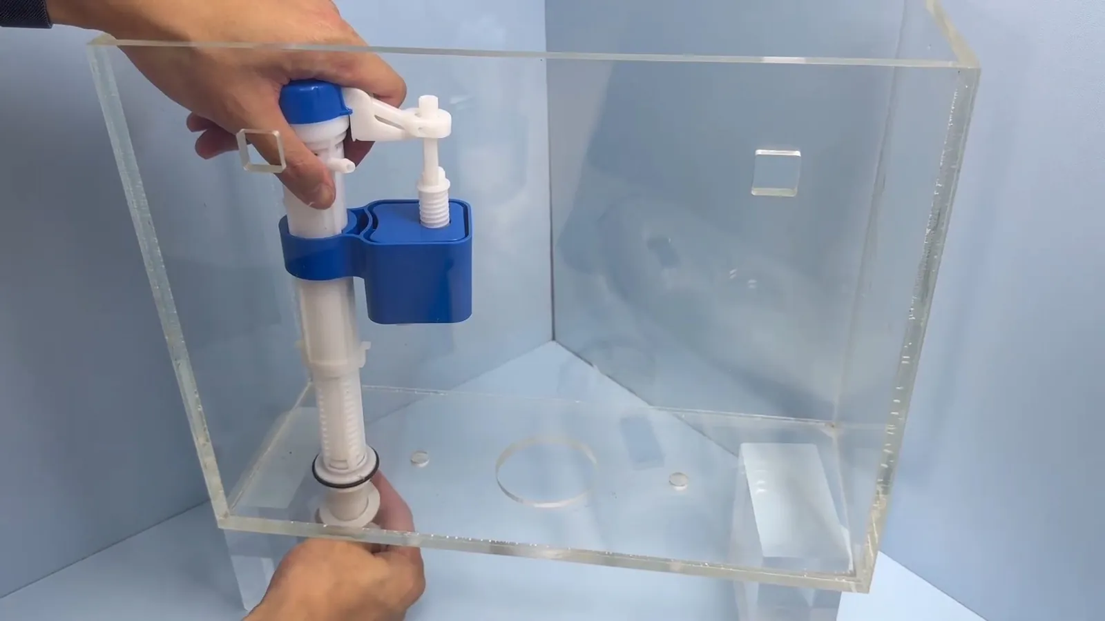 Hands adjusting a blue and white fill valve inside a clear acrylic toilet tank.