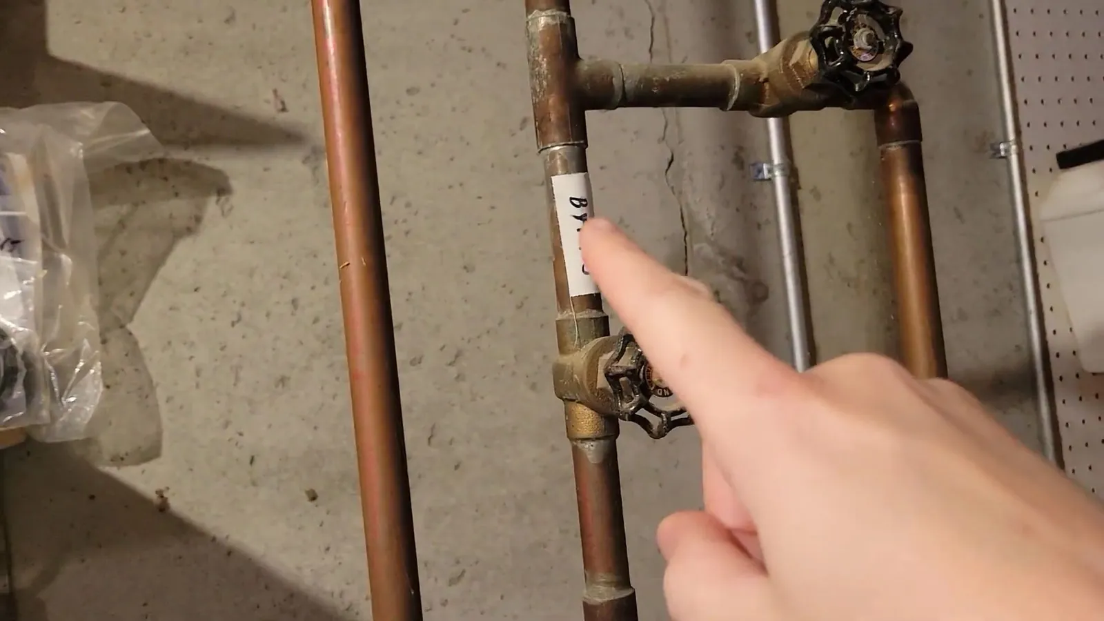Finger pointing at a brass shutoff valve between two copper pipes on a basement wall.