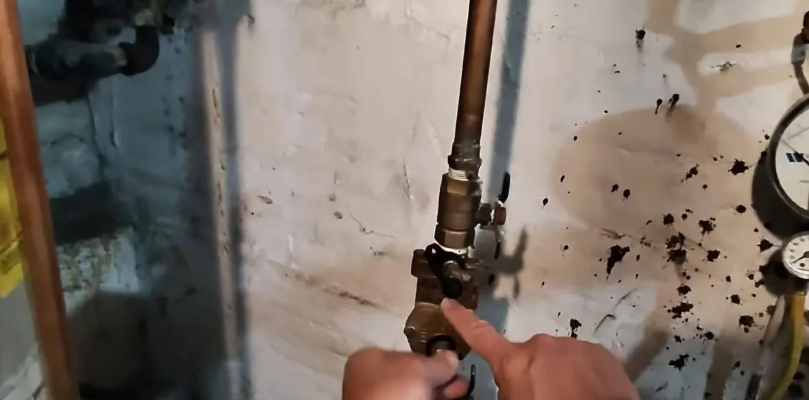 Finger pointing at a brass valve on a pipe beside a water meter dial with dark spatter on the wall.