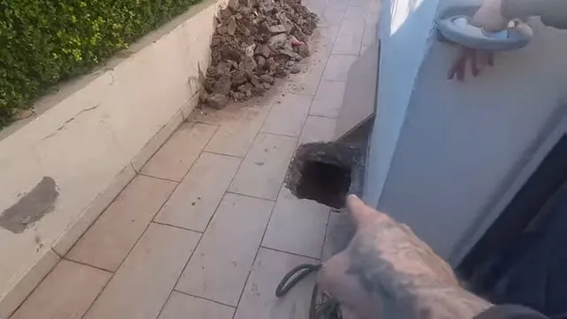 Tiled floor with a dark open cleanout hole beside a wall and a finger pointing to it.