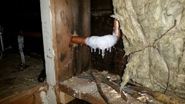 Copper pipe elbow wrapped in white ice with icicles hanging beside torn insulation in a wall cavity.