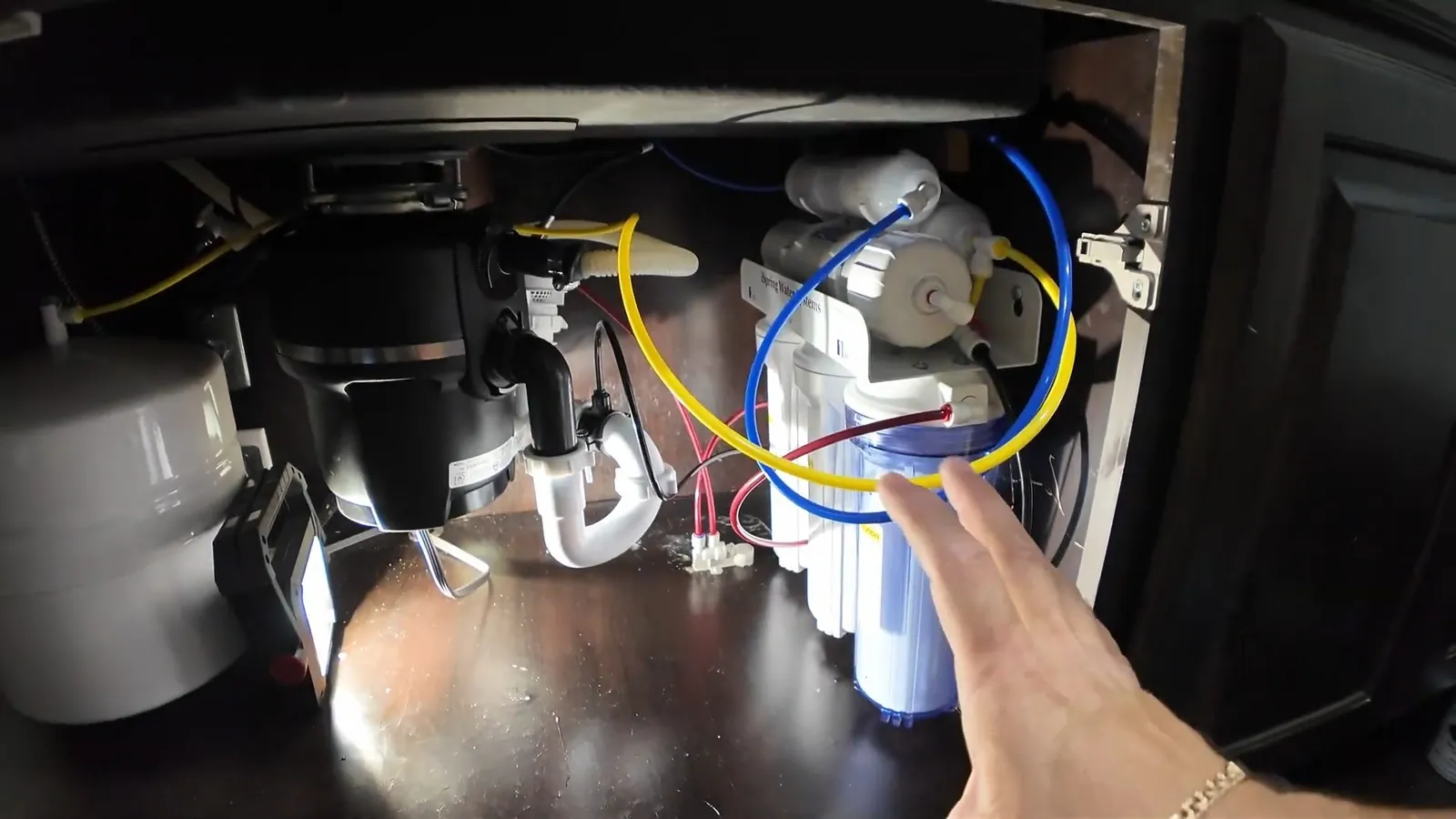 Open under-sink cabinet showing a garbage disposal beside a water filter and a hand touching a tube.