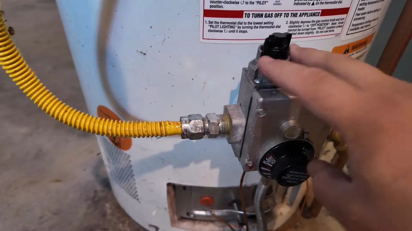 Hand on the gas control of a white tank water heater with a yellow gas line attached.