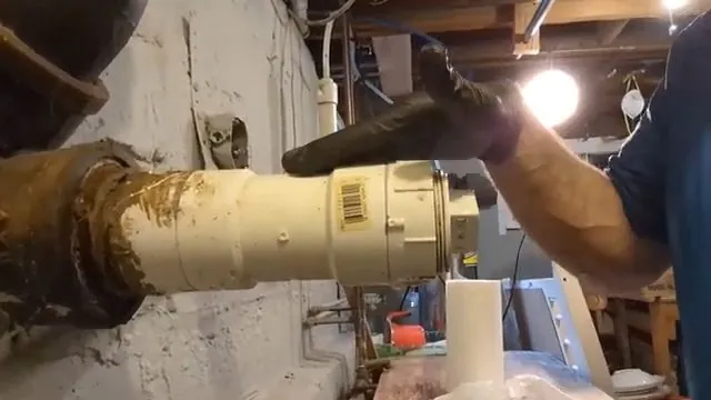 Gloved hand holding a cream-colored cleanout fitting with a barcode label on a drain line in a basement with concrete walls.