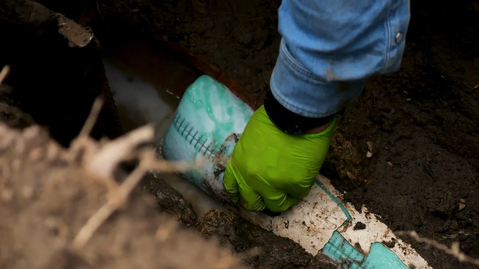 Hand in a green glove resting on a white pipe laid in a dirt trench.