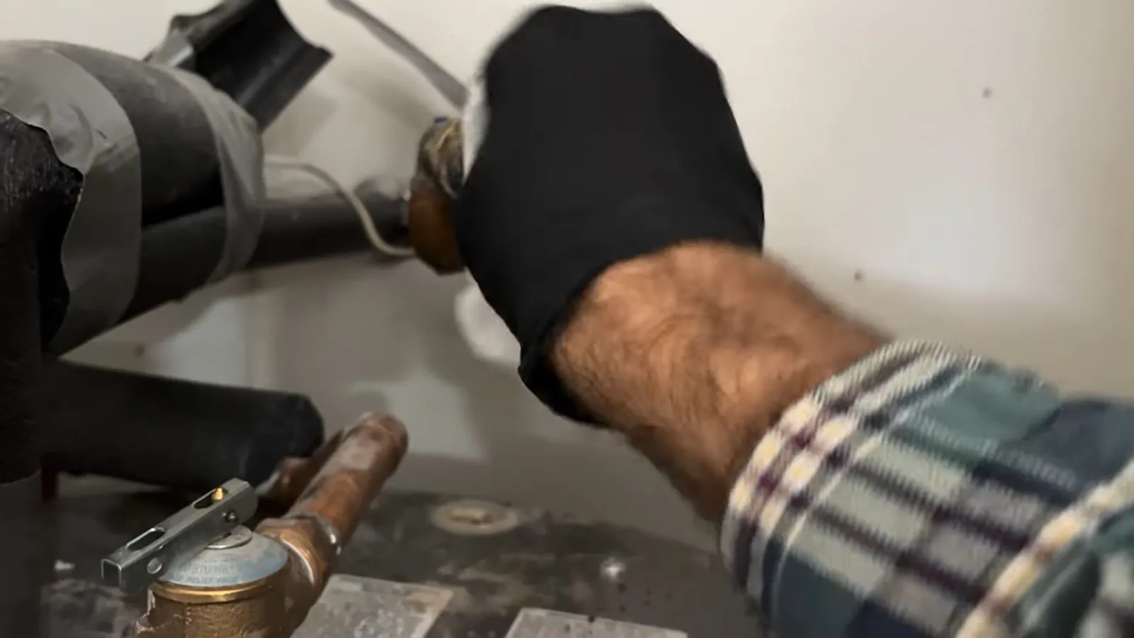 Gloved hand reaching to a pipe wrapped in black insulation above a water meter.
