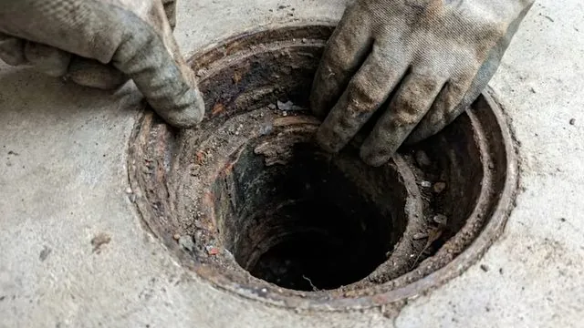 Gloved hands lifting the grate of a round floor drain set in a concrete floor.