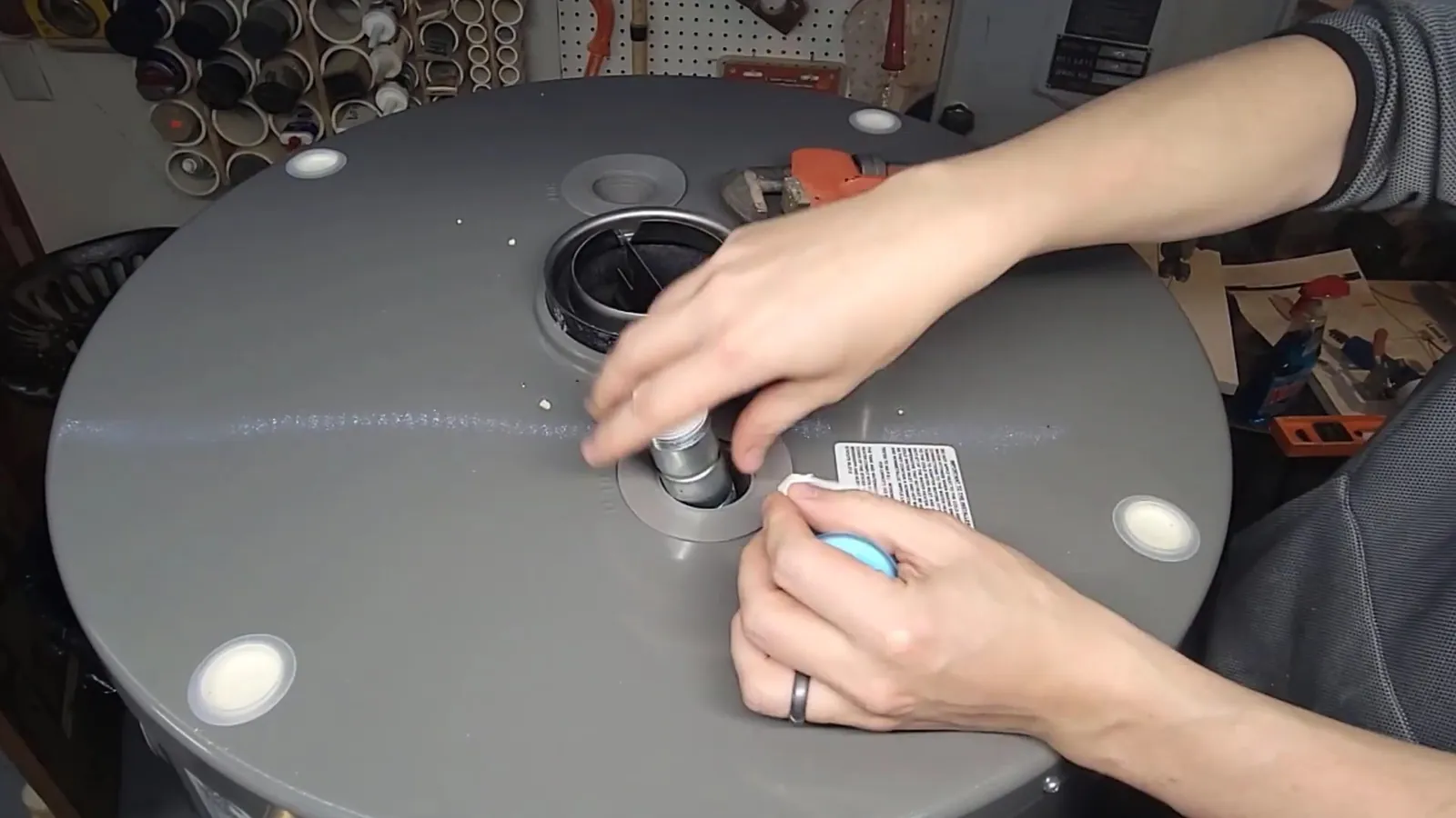Two hands working at a capped fitting on top of a gray tank water heater in a workshop.
