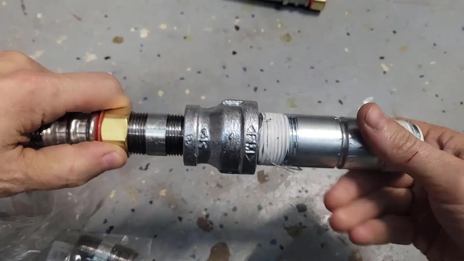 Two hands joining a threaded steel pipe union with a brass fitting on a speckled gray floor.
