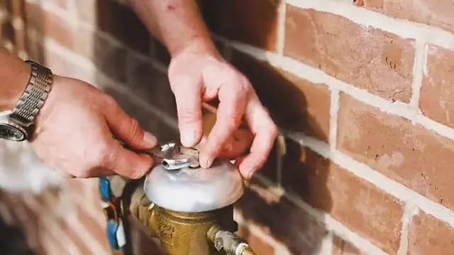 Hands lifting the cover of a brass water meter set against a brick wall.