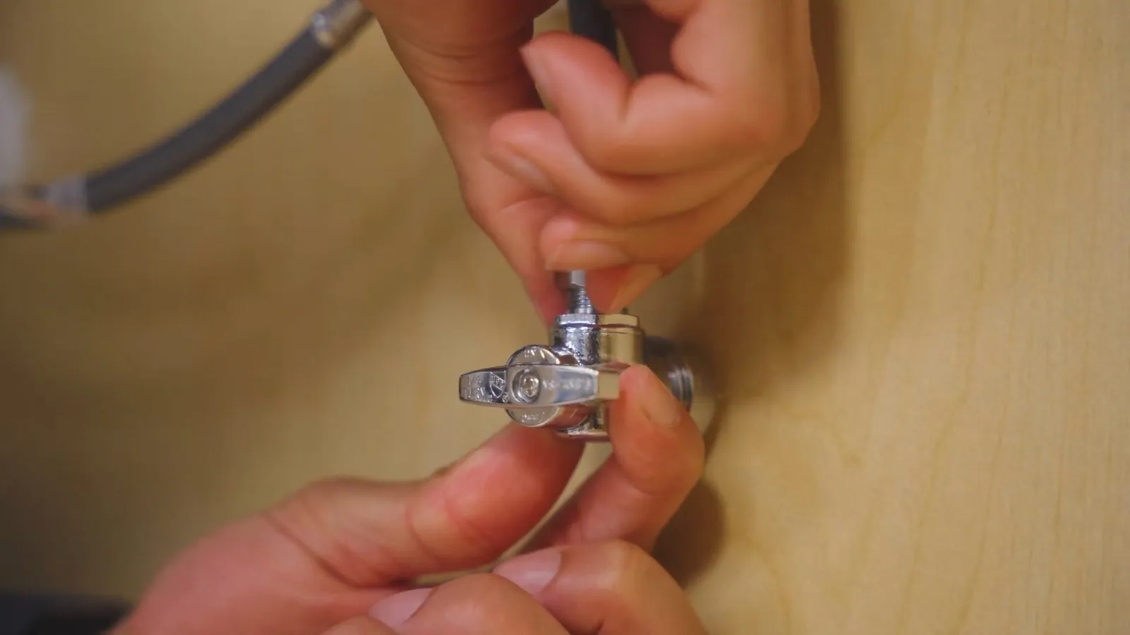 Two hands turning a chrome angle stop valve on the supply line under a sink.