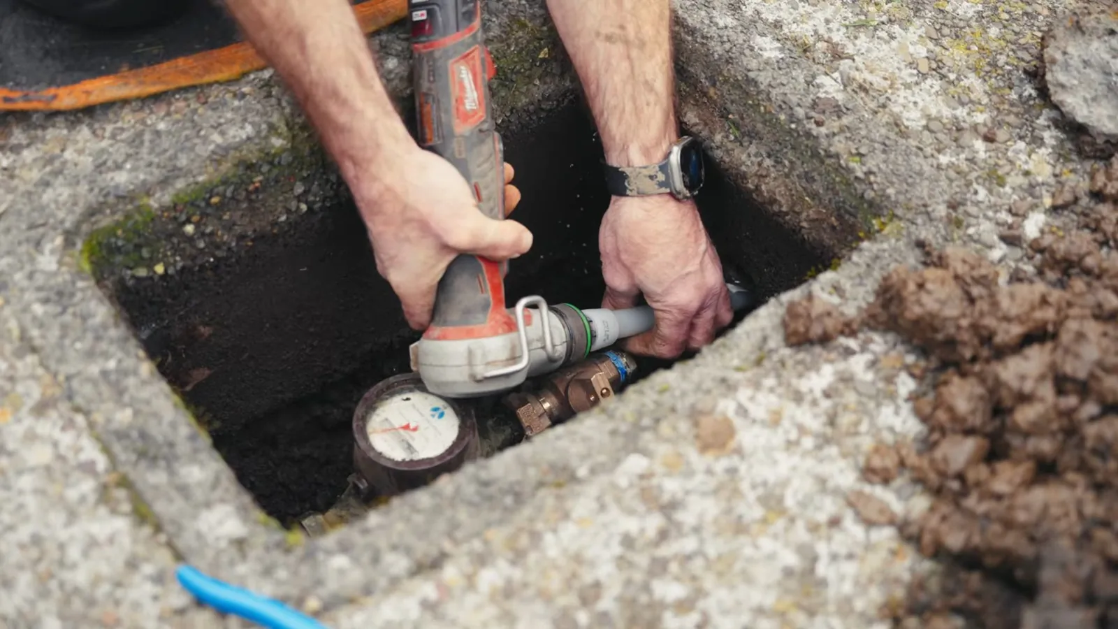Man's hands working with a power tool at a water meter inside a concrete ground box.