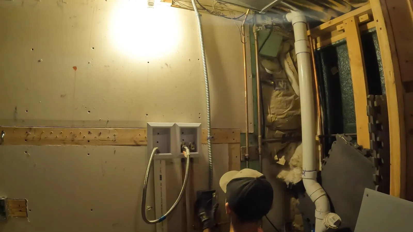 Man crouching in a basement beside white drain pipes and an open wall cavity under a bright work light.