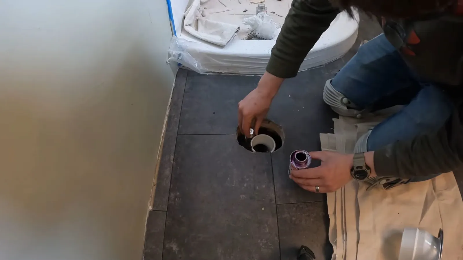 Man crouching on a bathroom floor over an open floor drain with a toilet behind him.