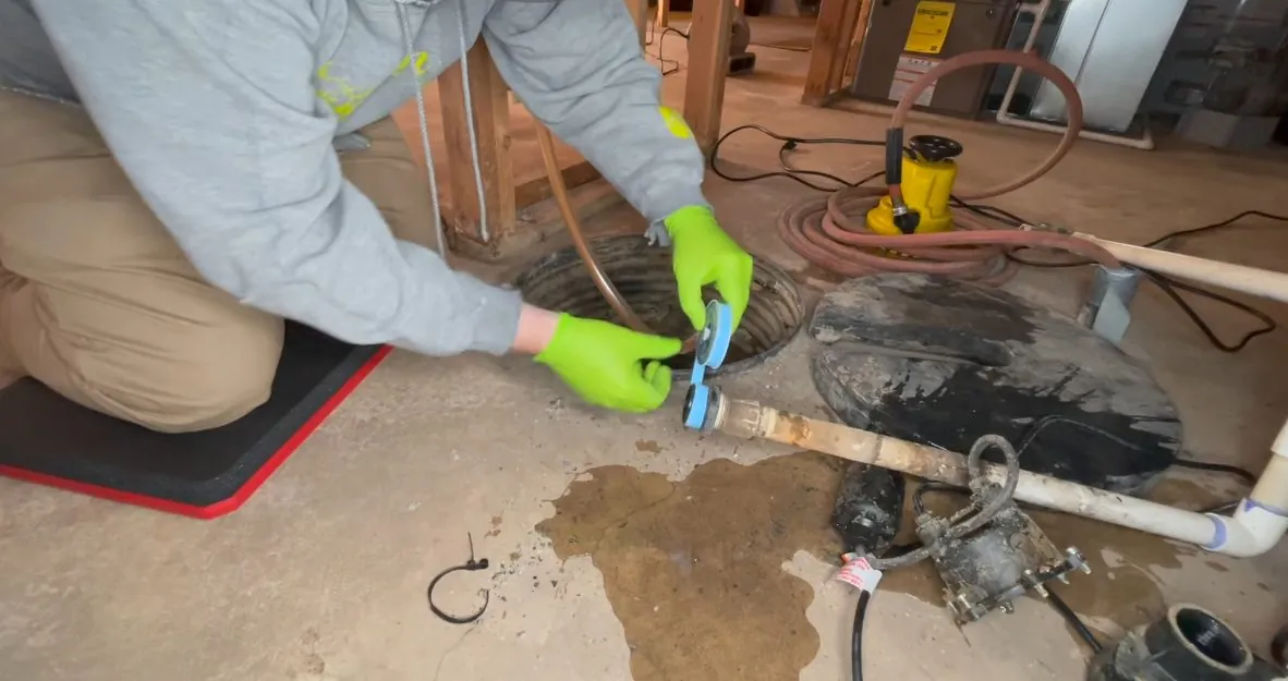 Man in gloves kneeling on a concrete floor beside a cut opening and a spreading wet patch.