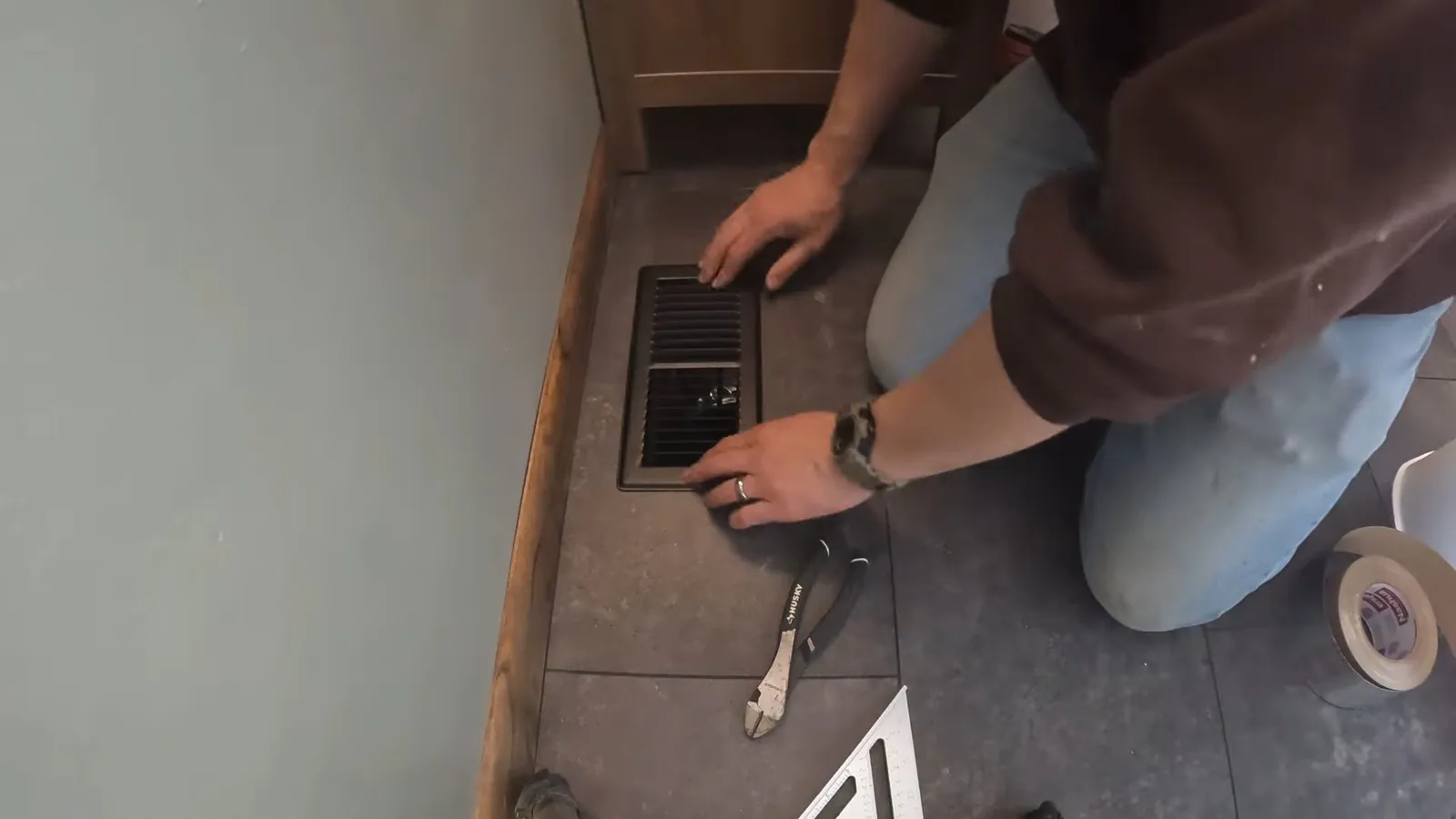 Man kneeling at a floor drain grate in a tiled corner with pliers and tape beside him.
