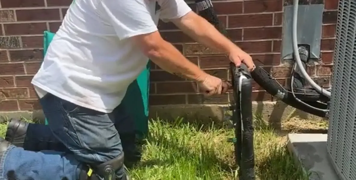 Man in a white shirt kneeling in grass beside a black cleanout pipe next to an outdoor unit.