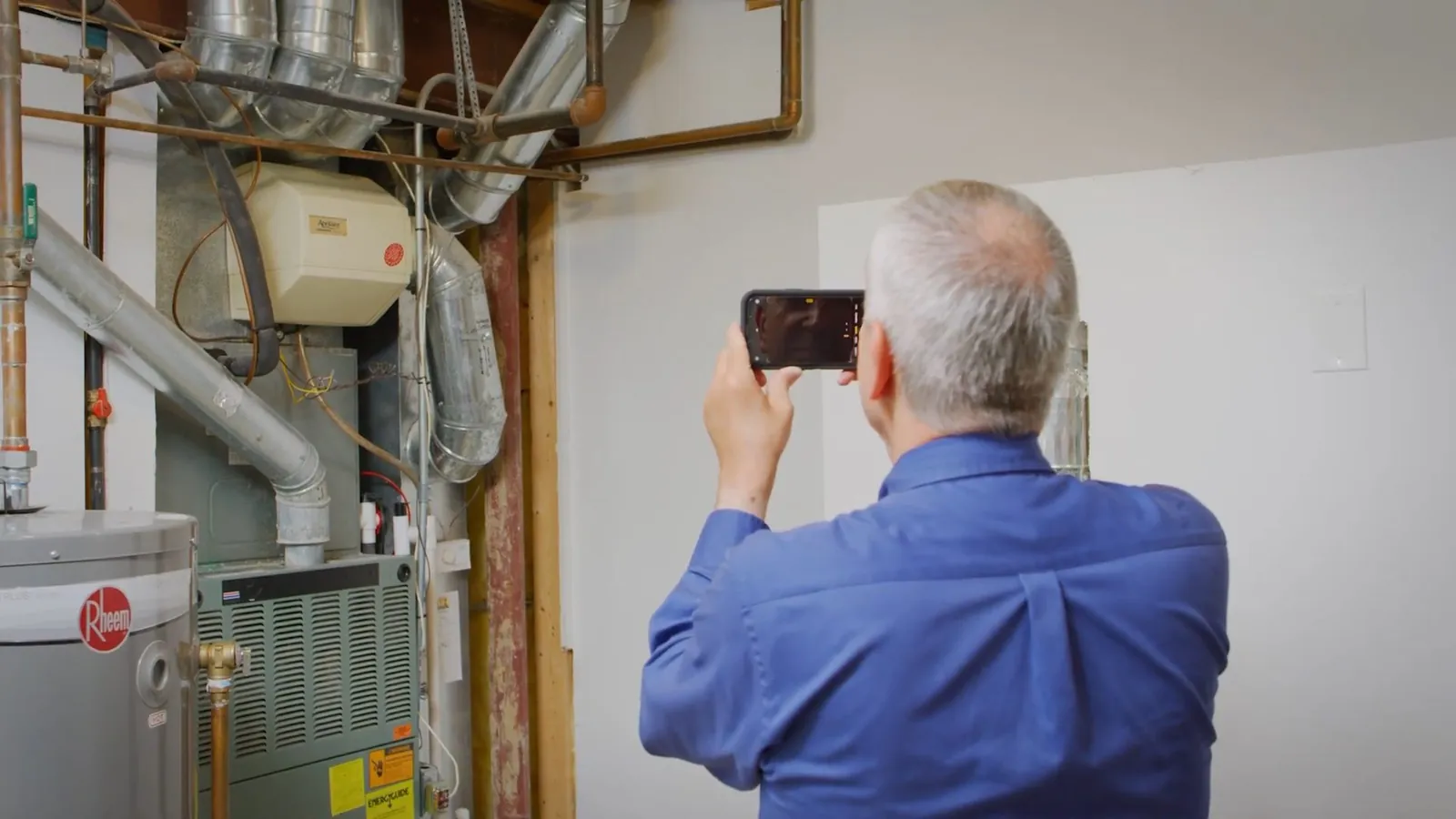 Man in a blue shirt photographing a gas water heater, furnace and pipes in a basement utility area.