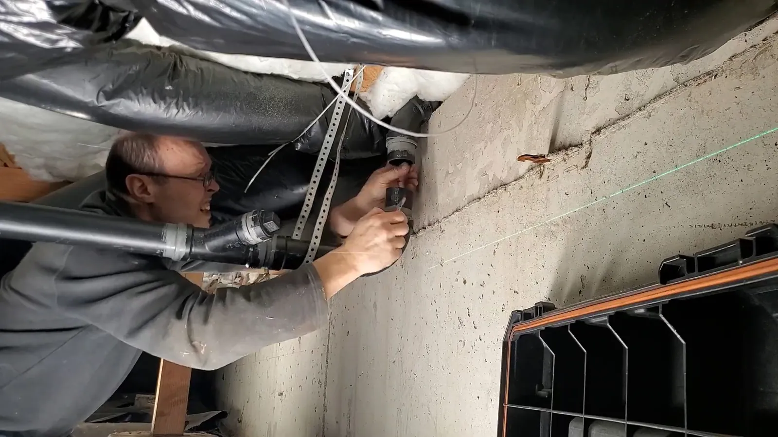 Man in a gray jacket in a basement reaching up to a pipe beside ductwork with a green laser line.