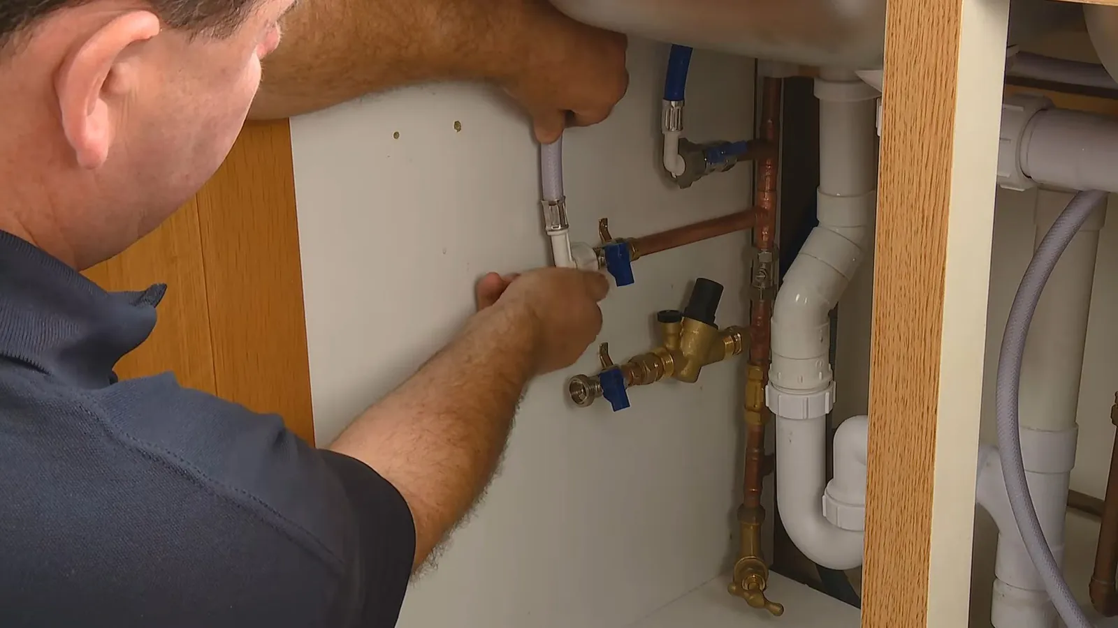 Man in a gray shirt reaching under a sink toward blue handled shutoff valves and white pipes.