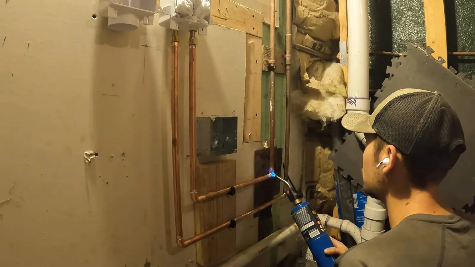 Man with a torch soldering copper pipe at a wall opening beside white drain pipes and insulation.