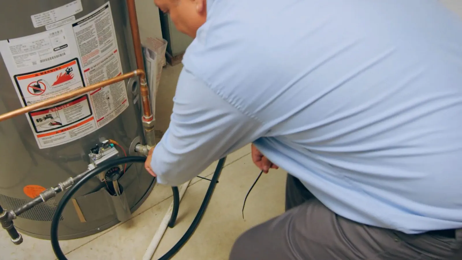 Man in a light blue shirt working on a gas tank water heater with its warning label visible.