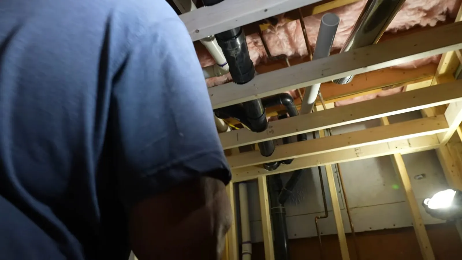 Man in a navy shirt working beneath exposed floor joists with black and white drain pipes overhead.