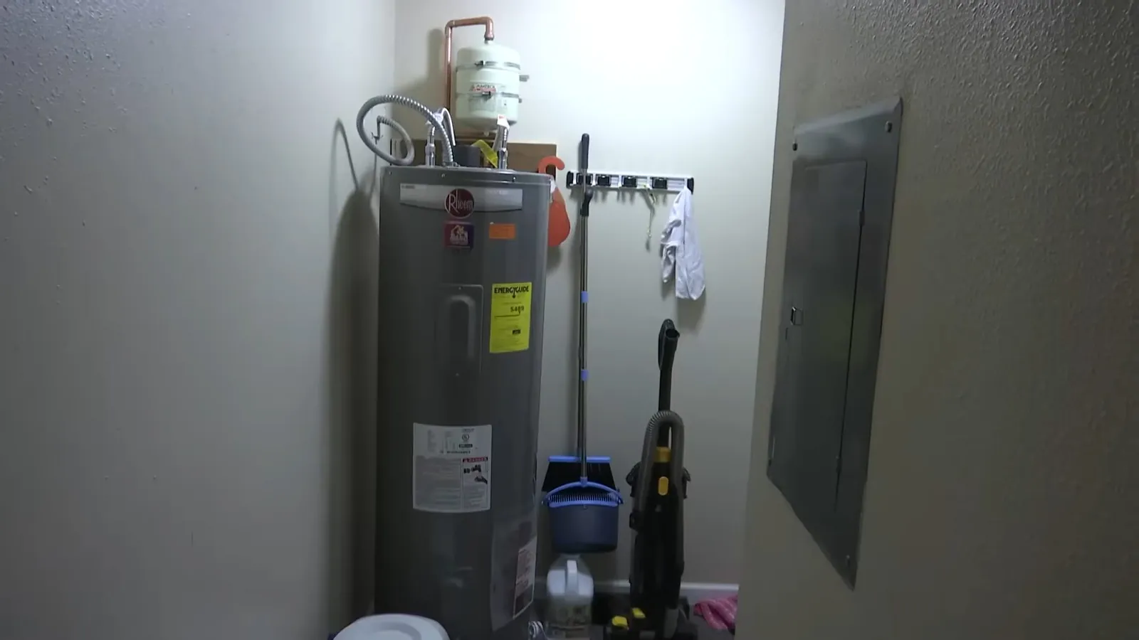 New tank water heater standing in a utility closet beside a panel and cleaning supplies.