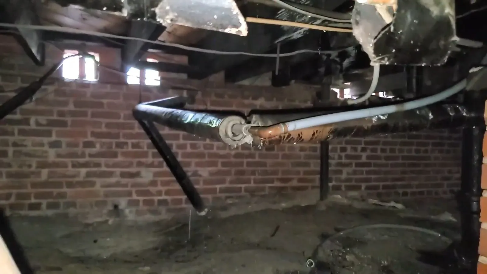 Older pipes running along a brick basement wall beneath exposed floor joists.