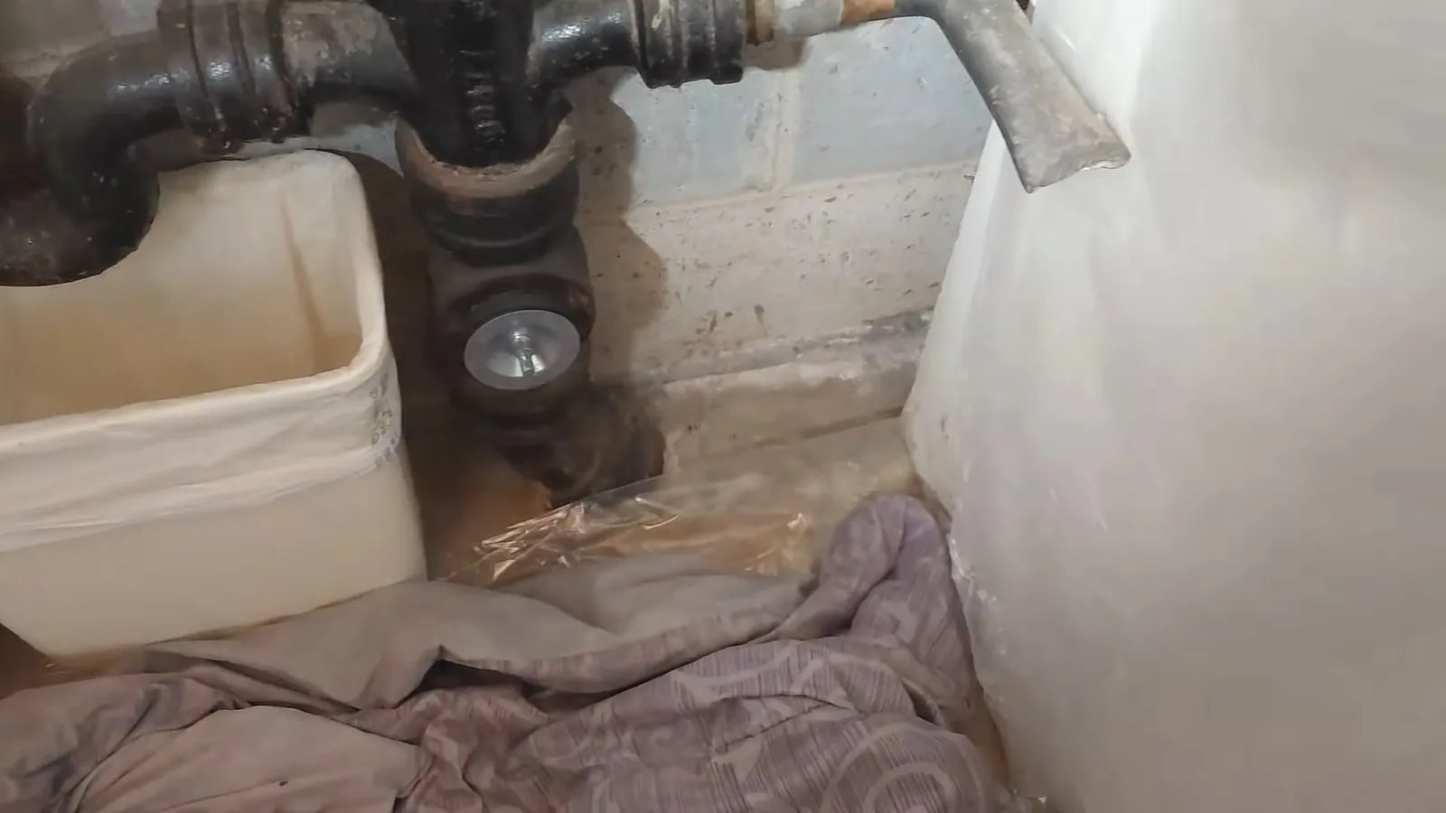 Old tank water heater beside black iron drain pipes, a white bin and towels on the floor.