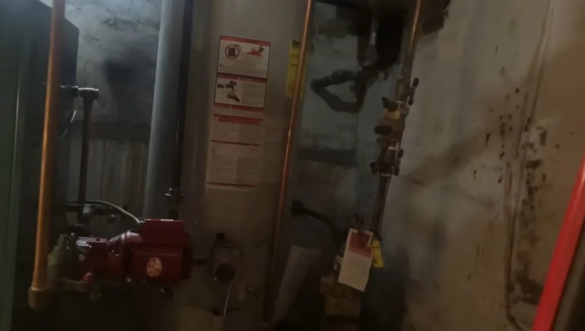 Older tank water heater in a basement with copper lines, a red valve and a warning label.