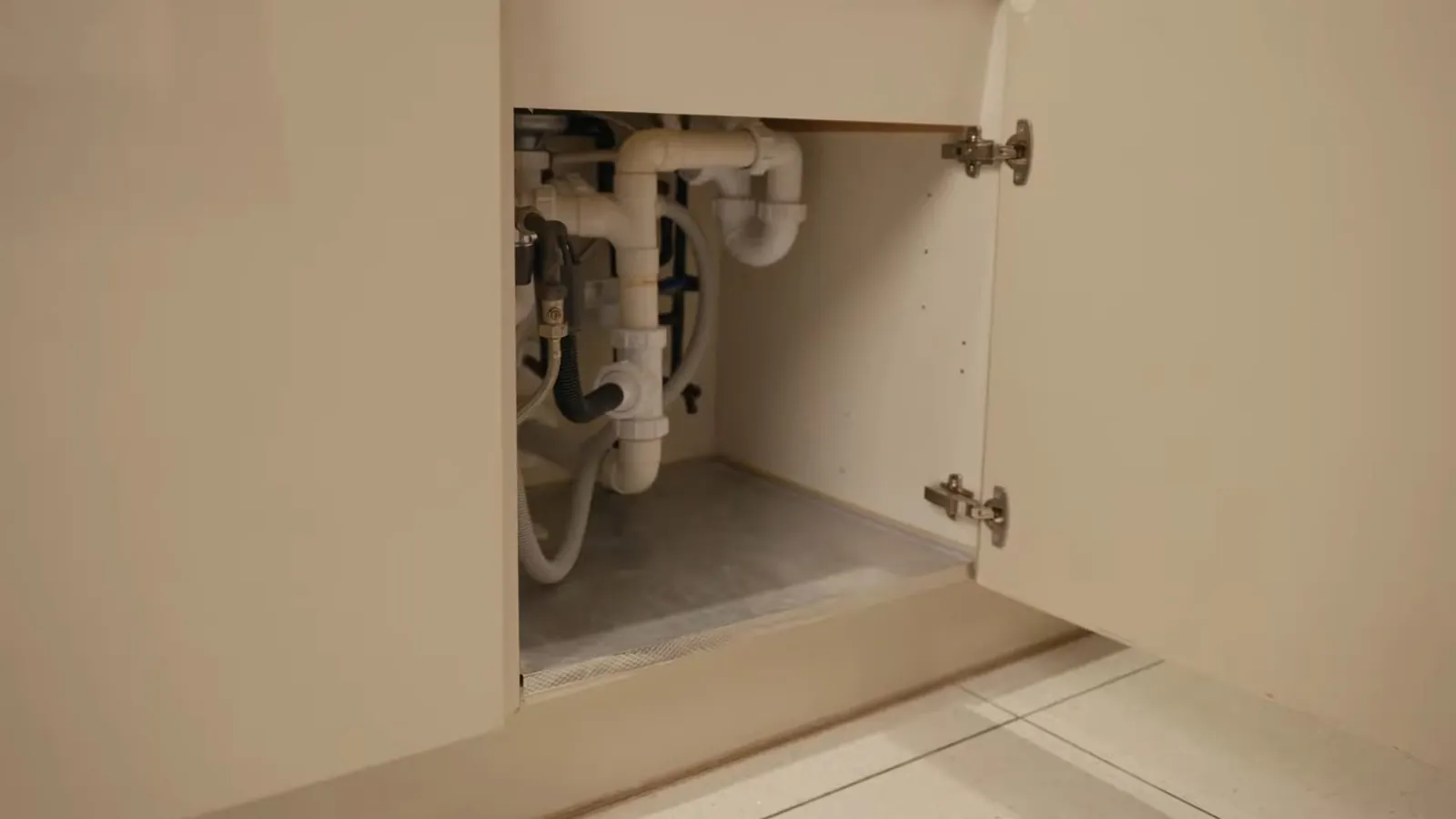 Open kitchen cabinet showing white drain pipes and supply lines below a sink.