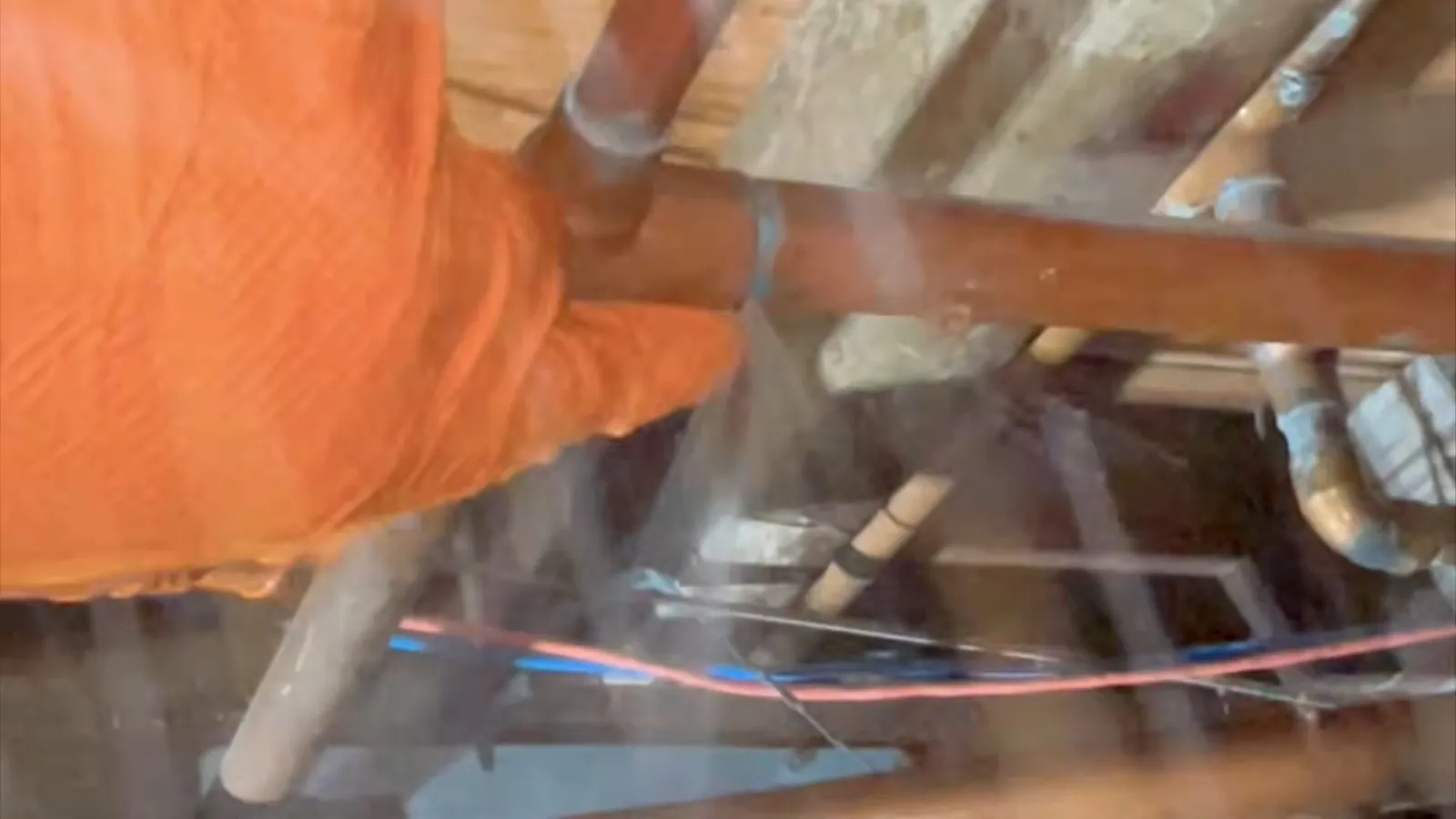 Orange-gloved hand beside water spraying from a pipe among floor joists.