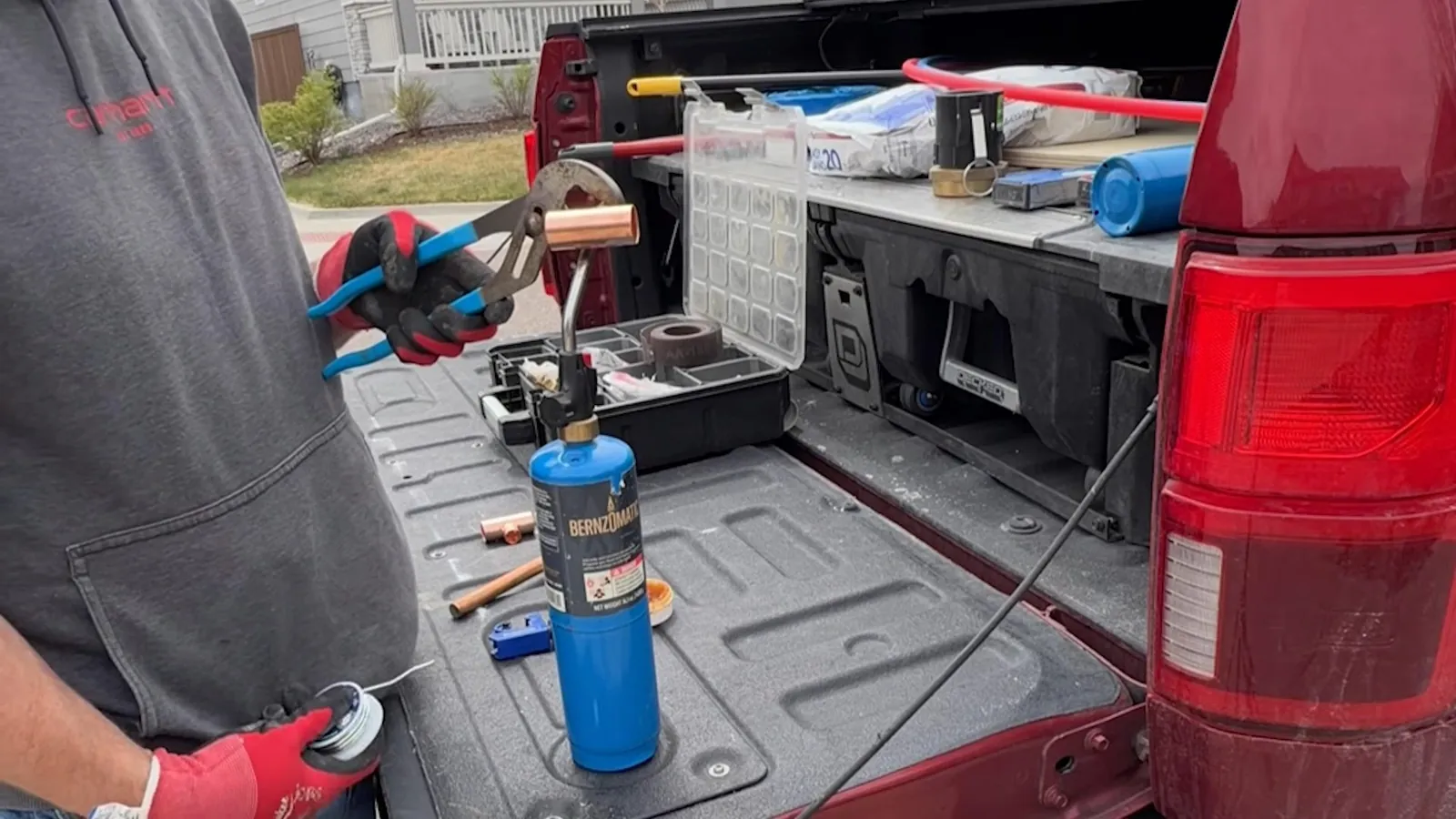 Open pickup truck bed holding a propane torch, tool trays and copper fittings, with houses behind it.