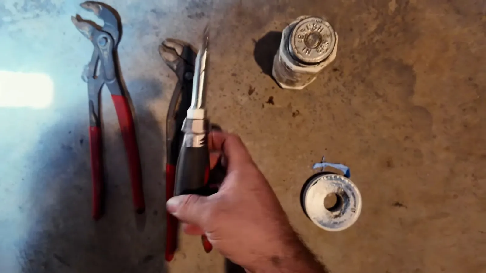 Pliers, a small pipe wrench and a spool of thread tape laid out beside a hand on a brown work surface.