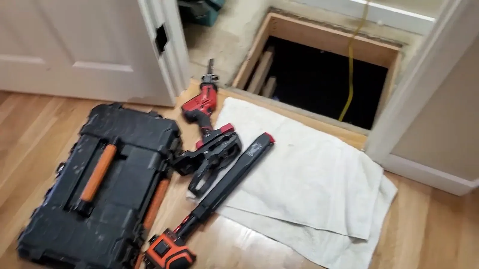 Open tool case, a drill and other tools on a white towel beside an open crawl space hatch in a wood floor.