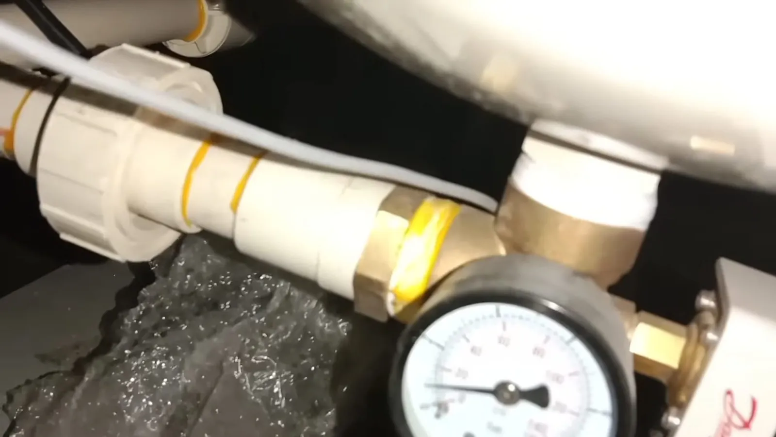 Pressure gauge and brass fitting on white pipe with water flowing beneath.