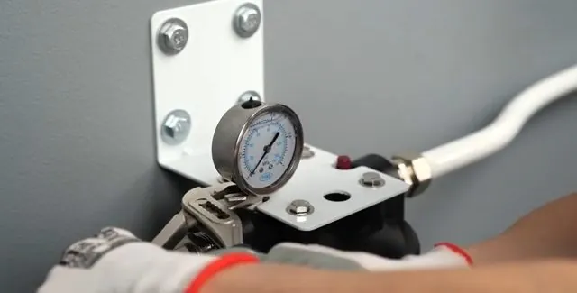 Pressure gauge on a bracket bolted to a wall with a white hose attached.