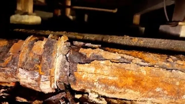 Heavily rusted and flaking cast iron pipe running under floor joists in a crawl space.