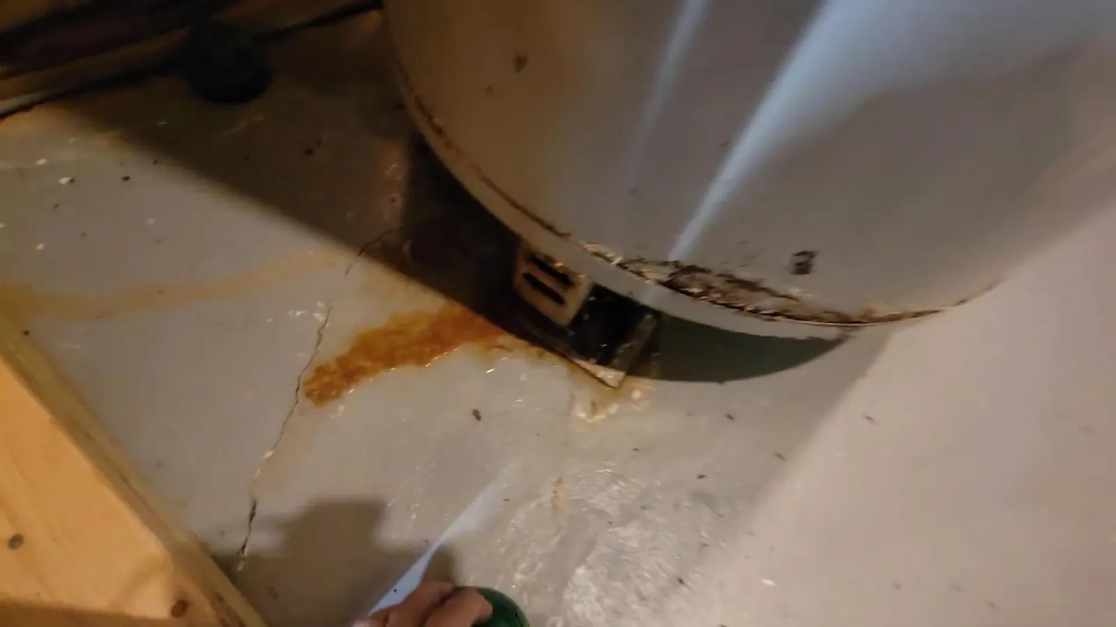 Base of a tank water heater with water spraying from it and rust staining the floor.